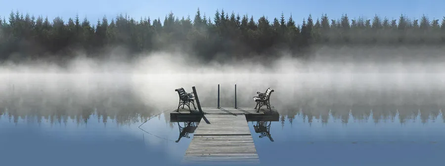 Brygga med två bänkar i morgondimma över sjön, med granskog i bakgrunden