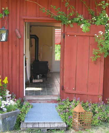 Öppen dörr i en röd stuga med blommor vid trappan